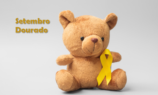 Foto de um ursinho de pelúcia marrom com um laço dourado em seu peito representando o setembro dourado, mês de conscientização do câncer infantojuvenil. Ao lado do ursinho o nome Setembro Dourado