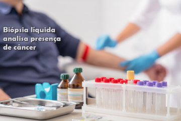 Imagem do braço de um homem e as mãos de uma enfermeira tirando sangue dele, em primeiro plano uma mesa com tubos para coleta do sangue que será enviado ao laboratório